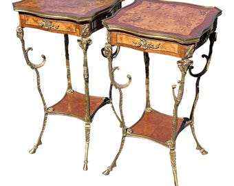 May include: Two antique side tables with ornate brass frames and burl wood tops. Each table features a small drawer with decorative hardware, a lower shelf, and curved legs. The tables have a classic, elegant design.