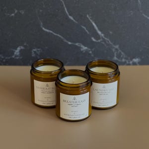 May include: Three amber glass candle jars with white labels. The labels have the names "Sanskrit Rose", "Breathe Easy", and "Laundry Day" printed on them. The candles are soy wax and hand-poured.