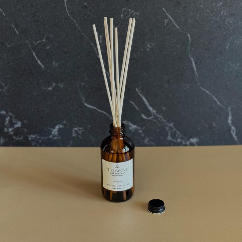 May include: A brown glass bottle with a white label that reads "THE GROVE" and "BLACKCURRANT & VANILLA". The bottle has a black lid and is filled with a clear liquid.  White diffuser sticks are in the bottle.