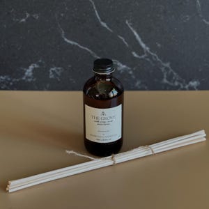 May include: A brown glass bottle with a black label that reads "THE GROVE" with the text "walk in orange and ginger" and "SOY WAX CANDLE OIL" on the label. The bottle is sitting on a brown surface with a bundle of white diffuser sticks.