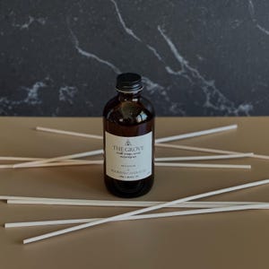 May include: A brown glass bottle with a black lid and a white label that reads "THE GROVE" with the text "wood orange, neroli, and vetiver" and "BOOKEND CANDLE CO.". The bottle is sitting on a brown surface with white sticks.