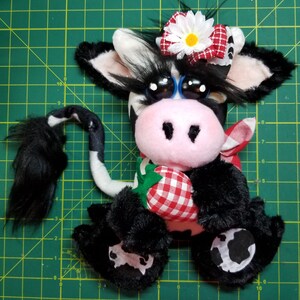 May include: A black and white plush cow toy with a pink nose and big blue eyes. The cow is wearing a red and white gingham bow and holding a red and white gingham strawberry.