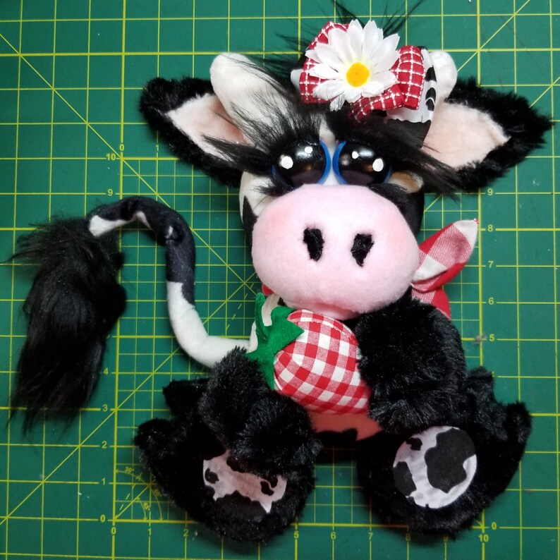 May include: A black and white plush cow toy with a pink nose and big blue eyes. The cow is wearing a red and white gingham bow and holding a red and white gingham strawberry.
