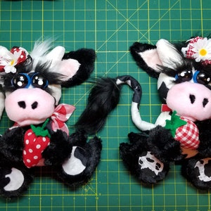 May include: Two black and white plush cow toys with large blue eyes, pink noses, and red and white gingham fabric accents. Each cow is holding a red and white strawberry with green leaves.
