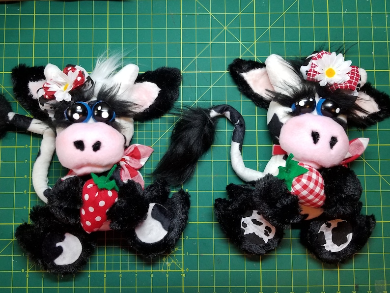 May include: Two black and white plush cow toys with large blue eyes, pink noses, and red and white gingham fabric accents. Each cow is holding a red and white strawberry with green leaves.