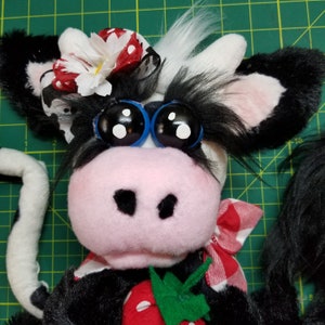 May include: A black and white plush cow with a red and white gingham bow tie, a red and white polka dot flower, and blue googly eyes. The cow is holding a green leaf and a red strawberry.