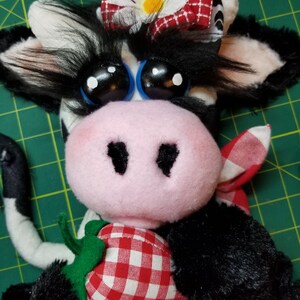 May include: A black and white plush cow with a pink nose and big blue eyes. The cow is wearing a red and white gingham bow with a white daisy on top. The cow is holding a red and white gingham strawberry.