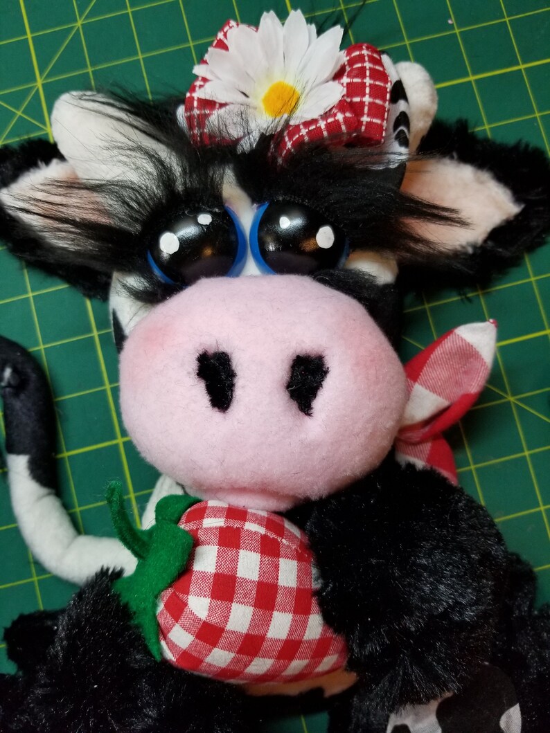 May include: A black and white plush cow with a pink nose and big blue eyes. The cow is wearing a red and white gingham bow with a white daisy on top. The cow is holding a red and white gingham strawberry.
