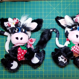 May include: Two black and white plush cow toys with big blue eyes, black eyelashes, and pink noses. Each cow is holding a red and white gingham strawberry. One cow has a white flower and red bow on its head, the other has a white daisy and red bow.