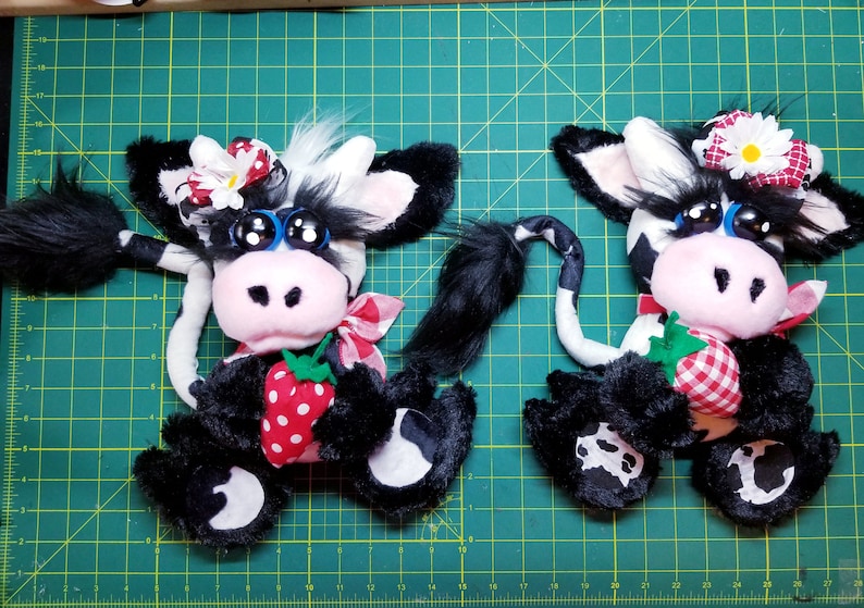May include: Two black and white plush cow toys with big blue eyes, black eyelashes, and pink noses. Each cow is holding a red and white gingham strawberry. One cow has a white flower and red bow on its head, the other has a white daisy and red bow.