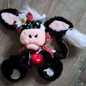 May include: A black and white plush cow toy with a red bandana, green eyes, and a white flower on its head. The cow is holding a red apple in its front paws.