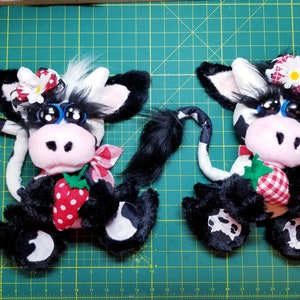 May include: Two black and white plush cow toys with pink noses and big blue eyes. Each cow is holding a red and white gingham strawberry. One cow has a white daisy on its head and the other has a red and white gingham bow.