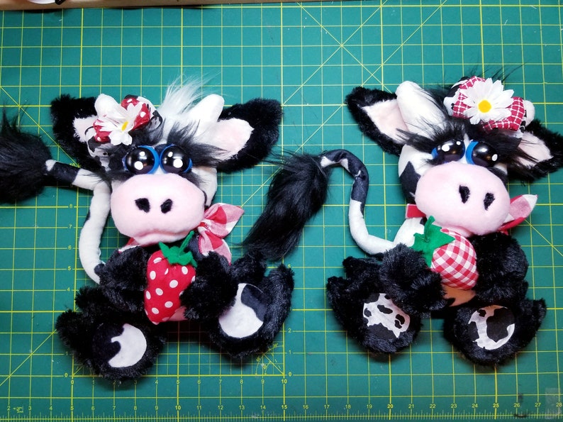 May include: Two black and white plush cow toys with pink noses and big blue eyes. Each cow is holding a red and white gingham strawberry. One cow has a white daisy on its head and the other has a red and white gingham bow.