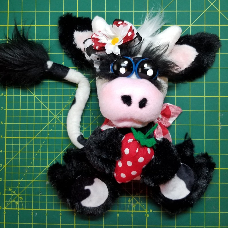 May include: A black and white plush cow with a red and white polka dot bow, red and white bandana, and blue sunglasses. The cow is holding a red and white strawberry in its front paws.