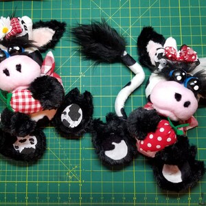 May include: Two black and white plush cow toys with red and white gingham fabric accents. Each cow is holding a red and white polka dot strawberry.