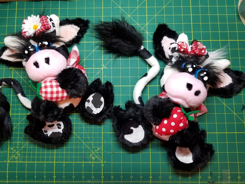May include: Two black and white plush cow toys with red and white gingham fabric accents. Each cow is holding a red and white polka dot strawberry.