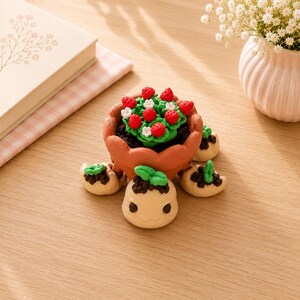 May include: A small turtle-shaped figurine with a terracotta-colored pot as its shell, containing a miniature green plant with red strawberries and white flowers. The turtle has a light beige head and four small beige feet with green leaves.
