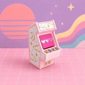 Può includere: Una statuetta di cabinato arcade in miniatura rosa e bianca con disegni celestiali dorati di lune, nuvole e stelle sul lato. Lo schermo mostra tre cuori bianchi pixelati su uno sfondo rosa brillante, con due piccoli joystick dorati.