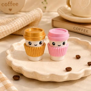 Op de afbeelding: Twee miniatuur koffiebekerfiguurtjes met lachende gezichtjes. De ene beker heeft een metallic gouden deksel en een geribbelde hoes, terwijl de andere een effen roze deksel en een bijpassende hoes heeft. Beide bekers zijn wit en staan op een schaal.