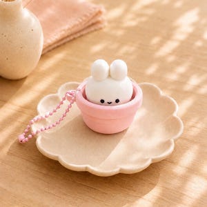 Könnte beinhalten: Eine kleine weiße Hasenfigur mit schwarzen Augen und einem winzigen Mund, die in einer hellrosa Tasse sitzt. Die Tasse verfügt über eine passende rosa Kugelkette an der Seite, wodurch sie als Schlüsselanhänger oder Anhänger verwendet werden kann.