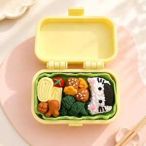 May include: A yellow plastic bento box containing miniature food replicas, including a white and black cat-shaped rice ball, green broccoli florets, orange meatballs, a red tomato, and a yellow rolled omelet, all resting on a green lettuce-like liner.
