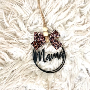 May include: Black wooden circle with the word "Mama" written in a cursive font. The circle is decorated with a brown and black leopard print bow and two wooden beads.