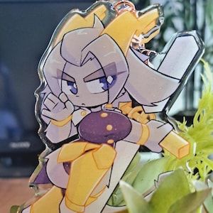 Könnte beinhalten: Ein klarer Acryl-Anhänger mit einer Cartoon-Figur in einer dynamischen Pose. Die Figur hat weiße Haare, blaue Augen und trägt eine gold-violette Rüstung. Der Anhänger ist vor einem Hintergrund aus grünen Pflanzen platziert.