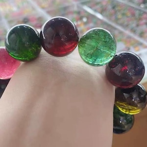 Puede incluir: Un brazalete con grandes cuentas de piedras preciosas pulidas en varios colores, incluyendo rosa, verde, rojo y negro. Las cuentas esféricas son translúcidas, mostrando patrones internos. El brazalete se muestra sobre un fondo claro.