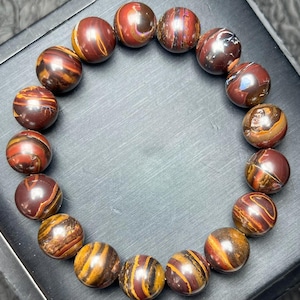 May include: A bracelet made of spherical beads in shades of red, brown, and gold. The beads have a striped pattern and are strung together. The bracelet is displayed on a dark surface. The beads are approximately 1 cm in diameter.