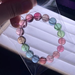 Puede incluir: Una pulsera de cuentas multicolores con cuentas redondas translúcidas. Las cuentas son en tonos rosa, verde, azul y marrón. La pulsera se sostiene en una bolsa de plástico transparente.