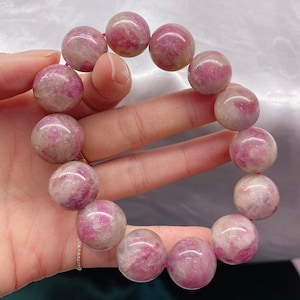 Puede incluir: Una pulsera con grandes cuentas esféricas en tonos rosa y blanco. Las cuentas están ensartadas en un cordón rosa, formando un círculo. La pulsera se sostiene en una mano, resaltando los patrones naturales de las piedras.