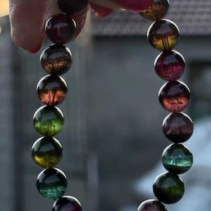 Puede incluir: Un brazalete de cuentas compuesto por esferas de piedras preciosas pulidas y multicolores. Las cuentas muestran una gama de colores, incluyendo rojos profundos, verdes y amarillos. El brazalete se sostiene, mostrando su forma circular y el juego de la luz en las piedras.