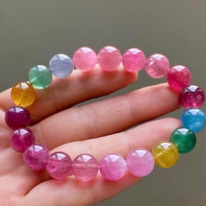 Puede incluir: Una colorida pulsera de cuentas con una variedad de piedras preciosas en tonos de rosa, morado, amarillo, verde y azul. Las cuentas redondas están ensartadas, creando un accesorio vibrante y llamativo. La pulsera se sostiene en una mano.