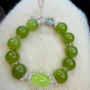 May include: A green jade bracelet with large, round beads and silver accents. The bracelet features a central oval jade stone surrounded by a decorative silver setting. A small silver charm hangs from the clasp.