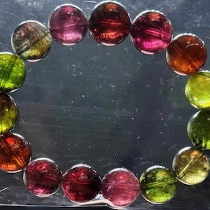 Puede incluir: Un brazalete hecho de piedras preciosas redondas y pulidas en tonos verdes, rojos y morados. Las cuentas están ensartadas, formando una forma circular. El brazalete se muestra sobre un fondo transparente y ligeramente reflectante.