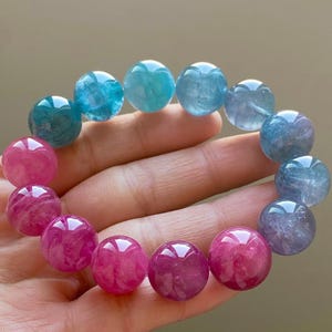 Puede incluir: Un brazalete hecho de cuentas redondas y lisas de piedras preciosas. Las cuentas son de un degradado de colores azul y rosa. El brazalete está en una mano.