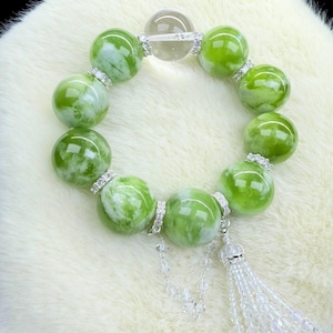 Könnte beinhalten: Ein Armband mit großen, runden, hellgrünen Perlen mit weißen Marmorierungen. Das Armband enthält eine klare, runde Perle und ist mit silberfarbenen Abstandshaltern und einer dekorativen Quaste versehen. Die Perlen haben einen Durchmesser von etwa 2,5 cm.