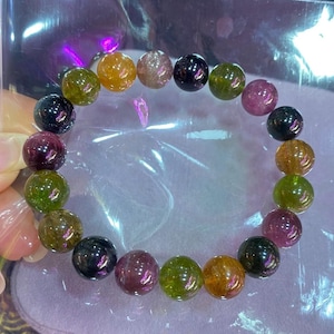 Puede incluir: Un brazalete con una cadena de cuentas de turmalina pulidas y multicolores. Las cuentas redondas muestran una variedad de colores, incluyendo verde, rosa, naranja y negro. El brazalete tiene una circunferencia de aproximadamente 18 cm.