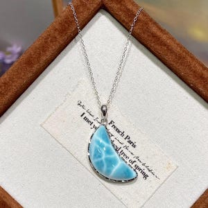 Könnte beinhalten: Ein halbmondförmiger Larimar-Anhänger in einer silberfarbenen Metallfassung, der an einer zarten silberfarbenen Kette hängt. Der Stein weist ein marmoriertes hellblaues und weißes Muster auf. Der Anhänger liegt auf einem weißen Stoffetikett.