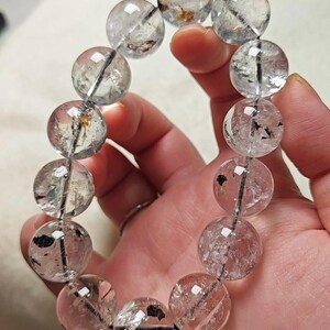 Peut inclure: Un bracelet composé de douze perles sphériques en quartz transparent. Chaque pierre présente des inclusions naturelles internes, notamment des taches minérales noires et brunes, enfilées sur un cordon fin, sombre et flexible visible à travers les perles.