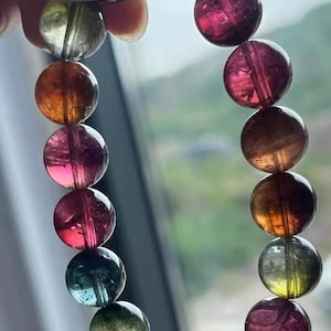 Puede incluir: Una pulsera de piedras preciosas multicolores con cuentas redondas en tonos rosa, naranja, verde y transparente. Las cuentas están ensartadas, creando un accesorio vibrante. La pulsera se sostiene sobre un fondo borroso.