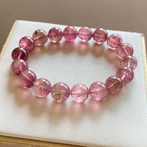 Puede incluir: Una pulsera de piedras preciosas rosas con cuentas redondas y translúcidas. Las cuentas, en tonos rosas y claros, están ensartadas. La pulsera se exhibe sobre una superficie blanca. Las cuentas miden aproximadamente 1 cm de diámetro.