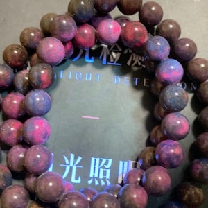 May include: A beaded bracelet featuring spherical stones with a mottled appearance in shades of deep purple, magenta, and dark blue. The beads are strung together in a circular arrangement, resting over a dark surface with text reading LIGHT DETECTION.