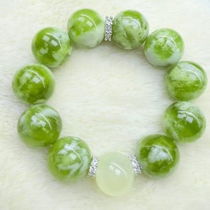 May include: A bracelet featuring large, round, glossy green beads with white marbling. The bracelet includes a light green oval bead and silver-colored connectors with small, clear stones. The beads are arranged on a white, fluffy surface.