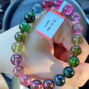 Puede incluir: Una pulsera de piedras preciosas multicolores con cuentas redondas en tonos rosa, verde, azul y marrón. Las cuentas tienen aproximadamente 10 mm de diámetro. Una etiqueta adjunta a la pulsera muestra el texto "era981" y "10mm 29.2g".