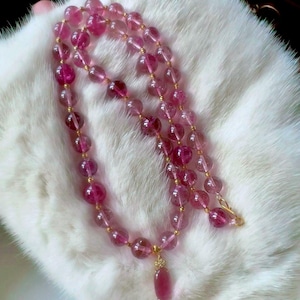 Puede incluir: Un collar de piedras preciosas rosas con un colgante. El collar presenta múltiples hilos de cuentas rosas redondas y translúcidas intercaladas con cuentas doradas. El colgante es una piedra preciosa rosa en forma de lágrima. El collar se muestra sobre una superficie blanca y esponjosa.