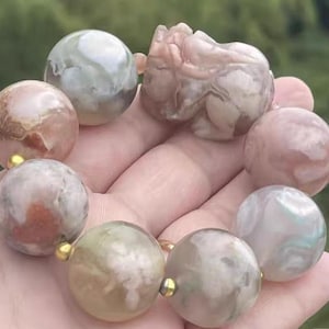 Puede incluir: Una pulsera con grandes cuentas redondas en tonos rosa, gris y crema, con una pieza central tallada. Las cuentas están ensartadas con detalles dorados, creando un accesorio único y elegante.