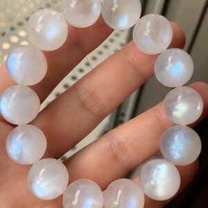 May include: A beaded bracelet composed of round, translucent white moonstone beads. Each bead exhibits a smooth, polished surface with a distinct iridescent blue sheen that reflects light, strung together on a clear, flexible elastic cord.