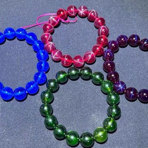 May include: Four circular bracelets made of smooth, polished spherical beads. Each bracelet features a distinct colour: one bright blue, one vibrant pink, one deep purple, and one dark green. Each bead displays a distinct four-pointed star light effect.