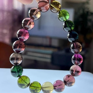 Puede incluir: Un brazalete compuesto por una serie de cuentas redondas, coloridas y translúcidas. Las cuentas son de color rosa, verde, amarillo y marrón. El brazalete se sostiene sobre un fondo claro.